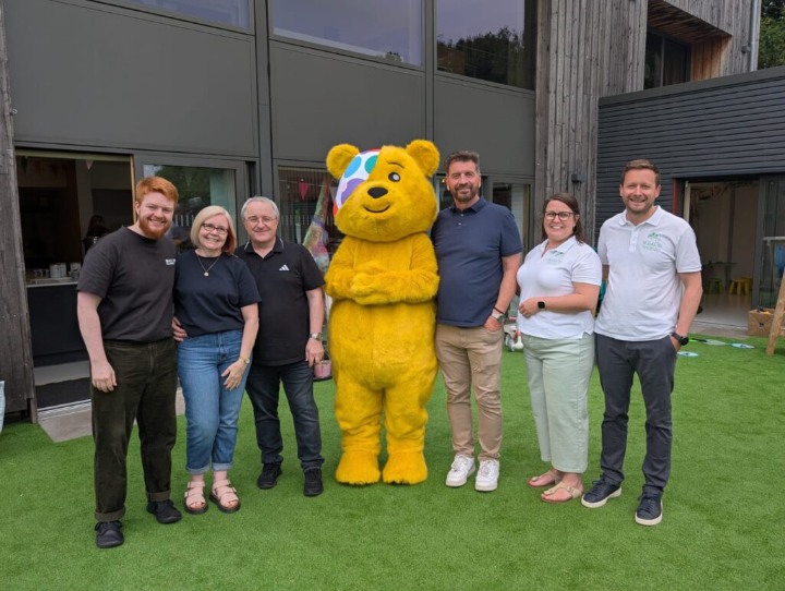 Group photo with Pudsey Bear mascot and six adults on a green turf, likely during a Children in Need charity event. Suitable for fundraising and corporate social responsibility.